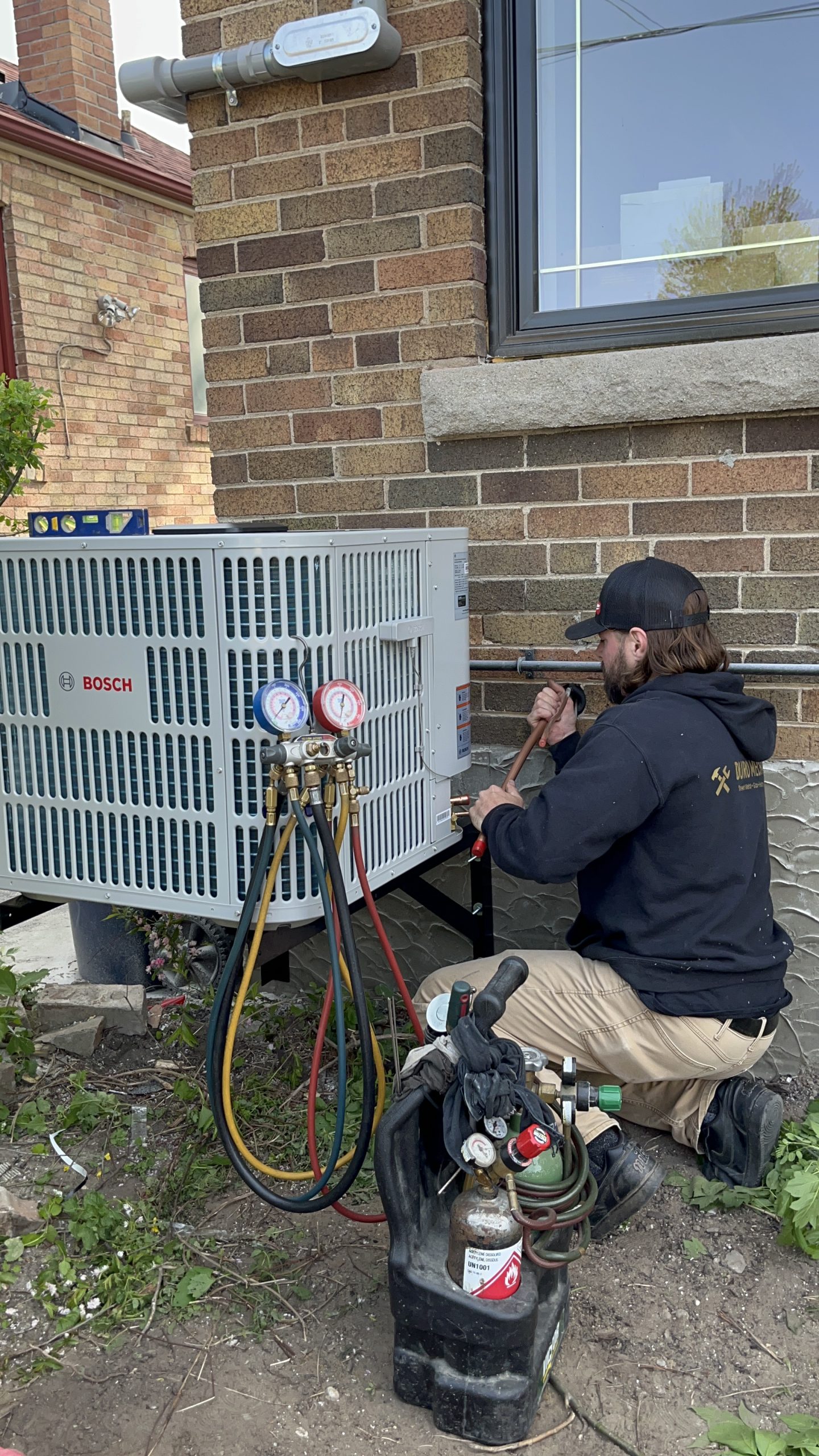 A technician working on an outdoor heat pump unit beside a house, performing maintenance or installation as part of an HVAC system.